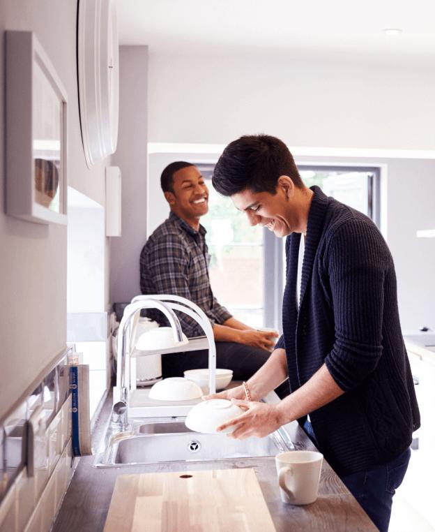 Two young men sharing a light-hearted moment in a bright, modern kitchen. The man in the foreground is washing dishes at the sink, wearing a dark sweater and smiling down at a white plate he's cleaning. The other man, wearing a plaid shirt, is seated at the kitchen island, leaning back and laughing. The natural light from the windows illuminates the kitchen, creating a relaxed and friendly atmosphere.