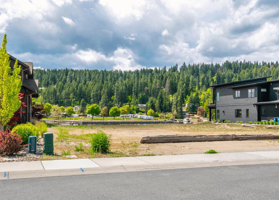 A residential street scene with modern houses on one side and a lush green forest in the background. The sky is partly cloudy, suggesting early summer weather. There is an empty, undeveloped lot in the foreground, with patches of grass and a large log lying across it. The houses have a contemporary design with dark and neutral colors, and the neighborhood appears quiet and newly developed.