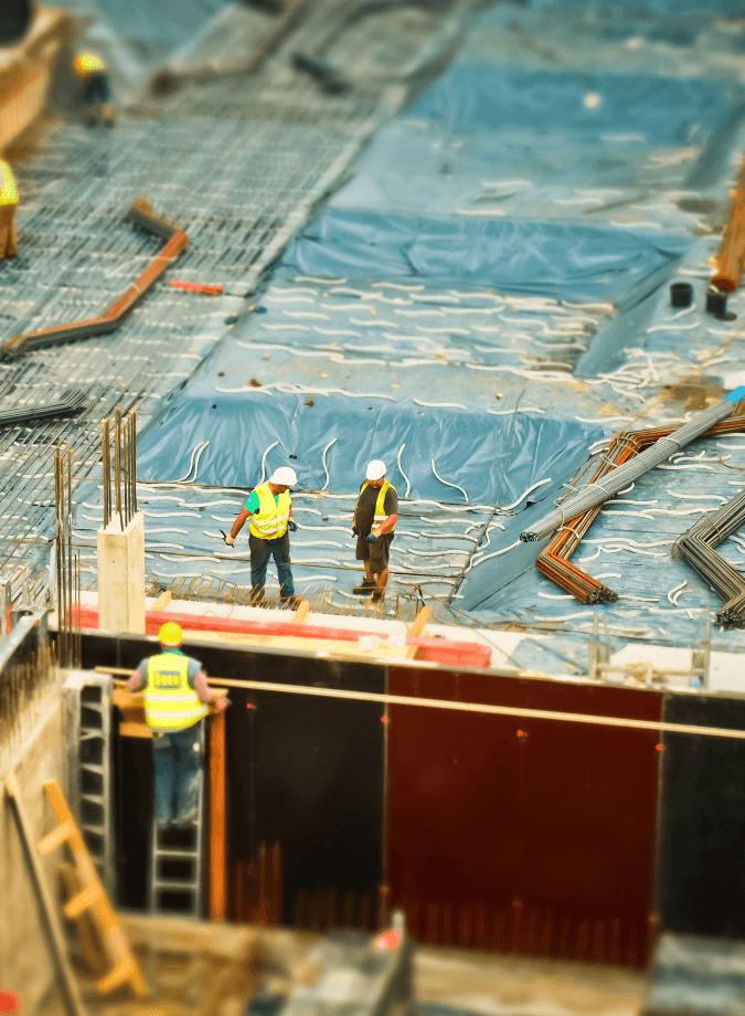 Construction workers on a building site. Three workers wearing bright safety vests and hard hats are standing on a concrete foundation with steel reinforcement bars. The site is covered with blue protective sheets, and there are piles of construction materials scattered around. The focus is on the workers, creating a miniature effect on the surroundings.