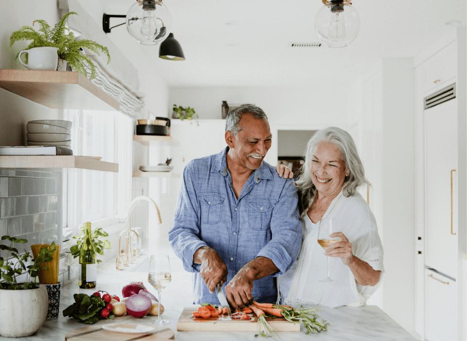An elderly couple is joyfully preparing food together in a bright, modern kitchen. The man, wearing a denim shirt, is slicing tomatoes on a wooden cutting board, while the woman, in a white blouse, is laughing and holding a glass of white wine. Fresh vegetables and a bottle of wine are on the countertop, with open shelving above adorned with plants and dishes. The atmosphere is warm and welcoming.