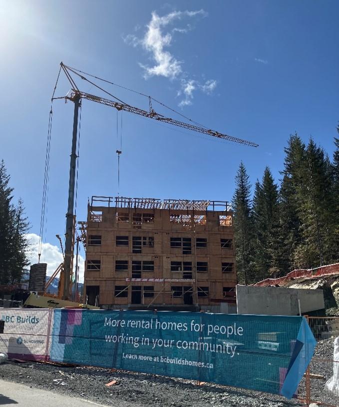 Construction site with crane and partially built wooden building, banner promoting rental homes, trees and blue sky in background.