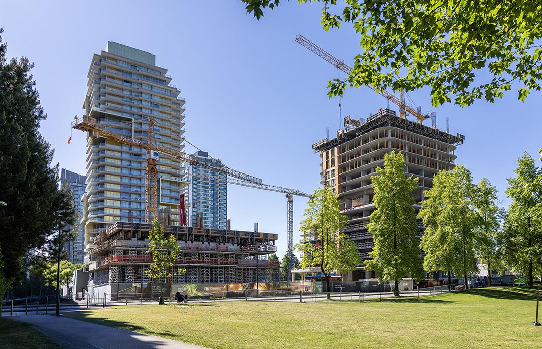 High-rise buildings under construction with cranes beside a green park and trees under a clear blue sky.
