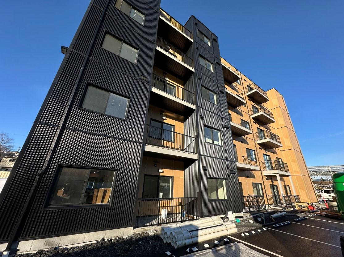 Modern apartment building with black and wood siding, balconies, and construction materials in a paved lot.